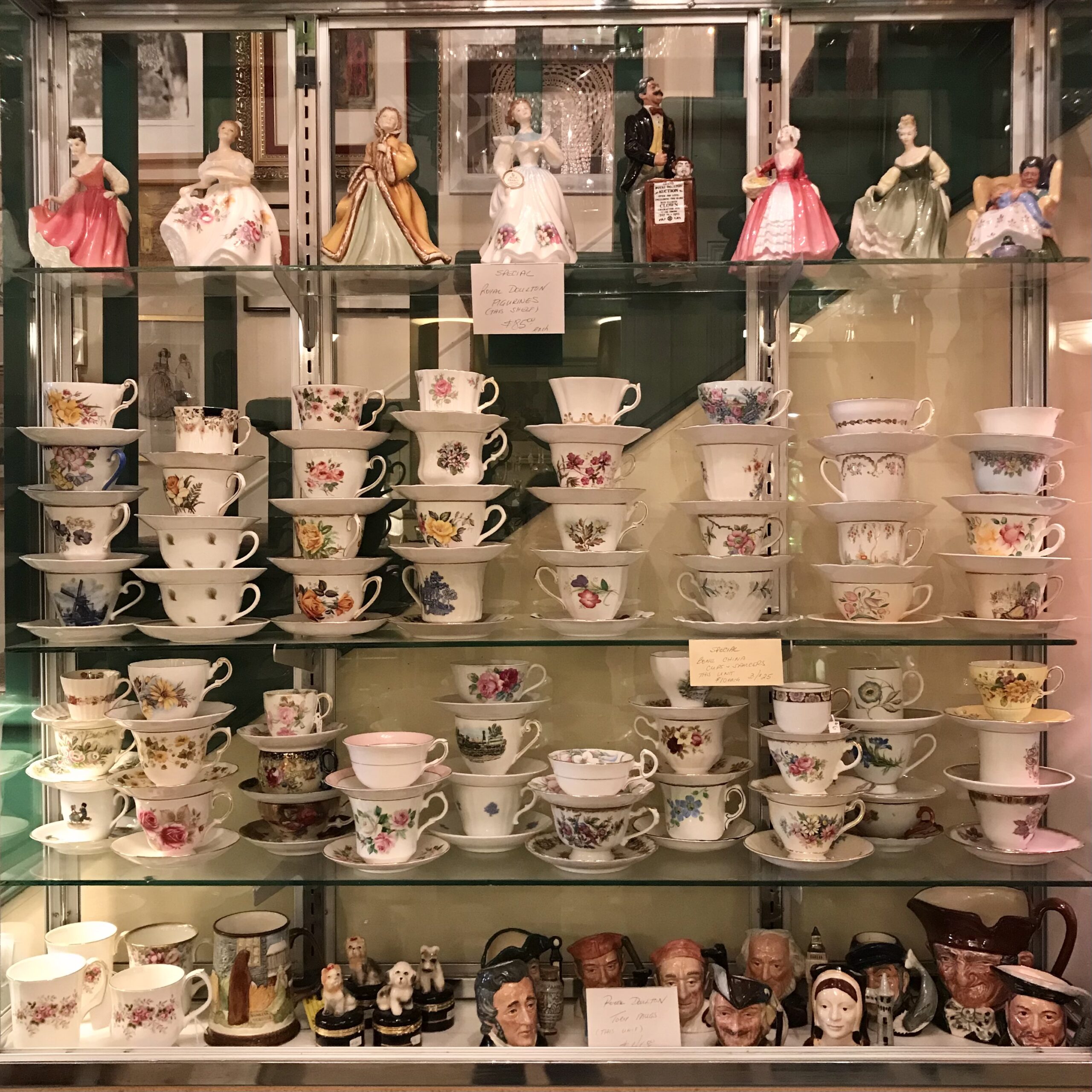 Four glass shelves holding Doulton figures at the top and cups and saucers on the lower shelves.